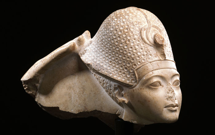 A statue fragment. On the left, part of a hand is visible, reaching out to touch the helmet worn by a young boy.  The nose of the boy has been broken away.