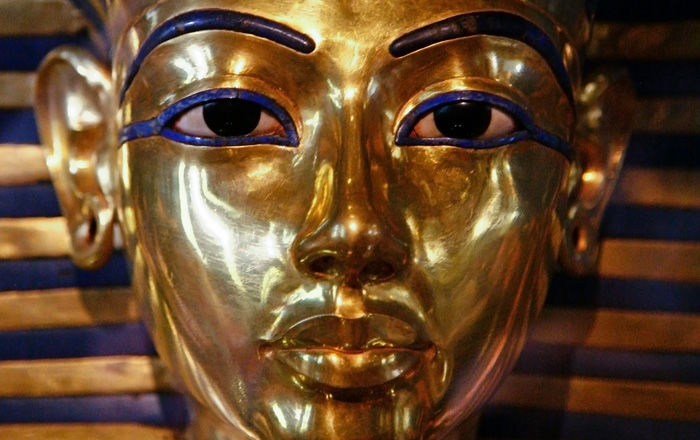 A close view of the face of the mask of Tutankhamun. It is gold, with the eyebrows and the lines around the eyes inlaid in blue and the eyes inlaid in white and black. The gold and blue horizontal stripes of the royal headdress are visible to the sides of the face.
