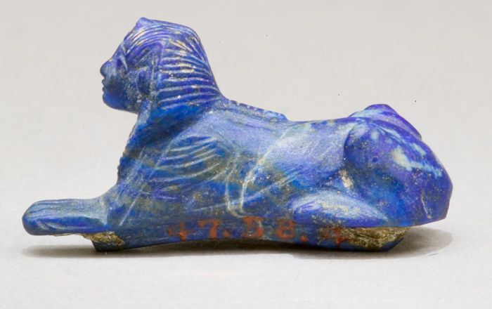 A deep blue sphinx, recumbent and facing left. An incised wing is visible on the visible shoulder of the lion, the human head wears a striped headdress, with the stripes indicated with incisions.