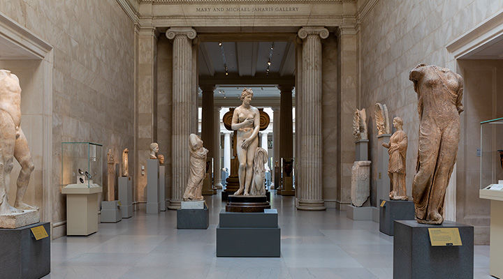 A marble statue of Aphrodite stands in the middle of the Greek and Roman hall surrounding by other statues and sculptures. She is standing upright with her hands moving to cover her breasts and pubic area. 