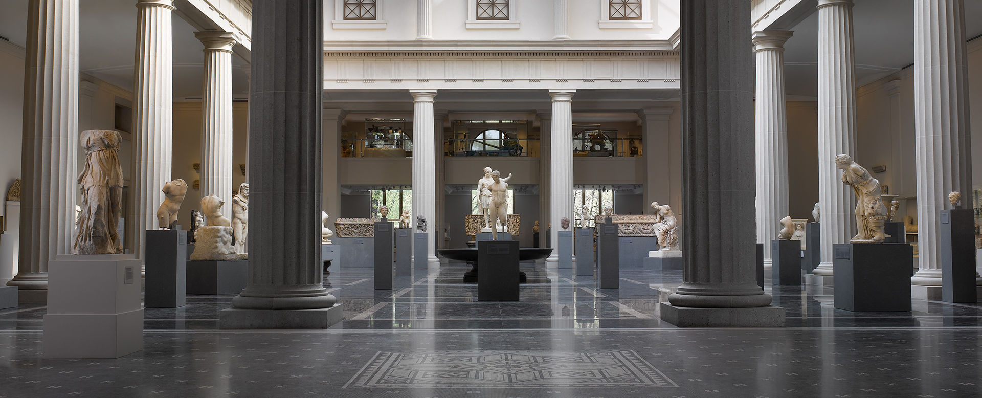 Una galleria molto ampia racchiusa da un colonnato e piena di sculture in pietra dell'antica Roma illuminate dalla luce solare naturale.