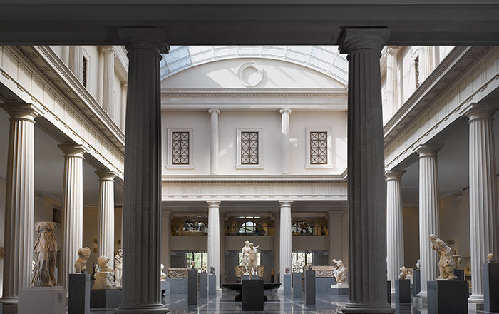 A very large gallery enclosed by a colonnade and filled with with stone sculptures from ancient Rome lit by natural sunlight.