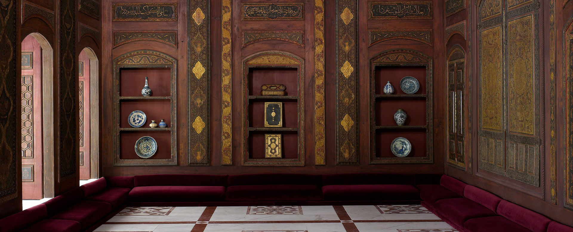A close-up of a room from Damascus decorated with gilded dark wood paneling, low dark red velvet covered cushions; open windows on the left illuminate the room with light