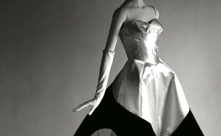 Cropped view of a mannequin against a grey, spotlit seamless background; the mannequin is dressed in long white evening gloves and a strapless, sweetheart white reflective gown with large black shape around the skirt.