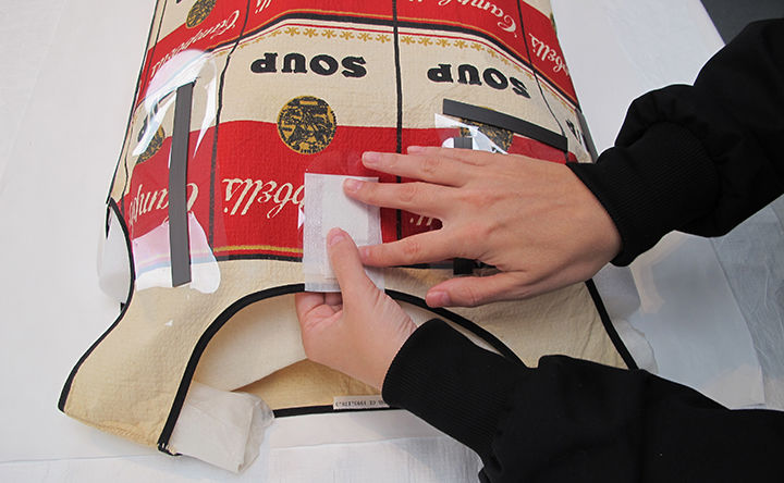 A pair of hands appears to tape a patch of material atop a sleeveless dress resting on a table surface; the dress is made out of a fabric patterns with Campbell's iconic red and white soup can package design