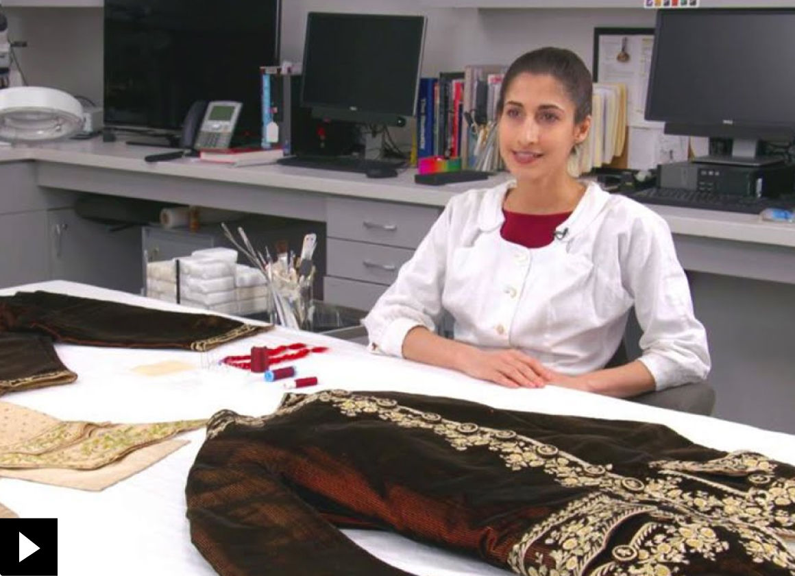 A conservator sits at a table where elements of an dark brown 18th century suit have been spread out across the table surface; there is a small play button icon in the bottom left-hand corner