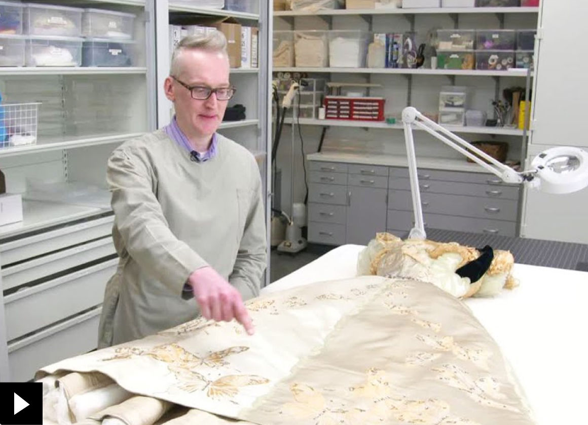 A conservator points to an area of an embroidered gown resting across a large white table; there is a small play button icon in the bottom left-hand corner
