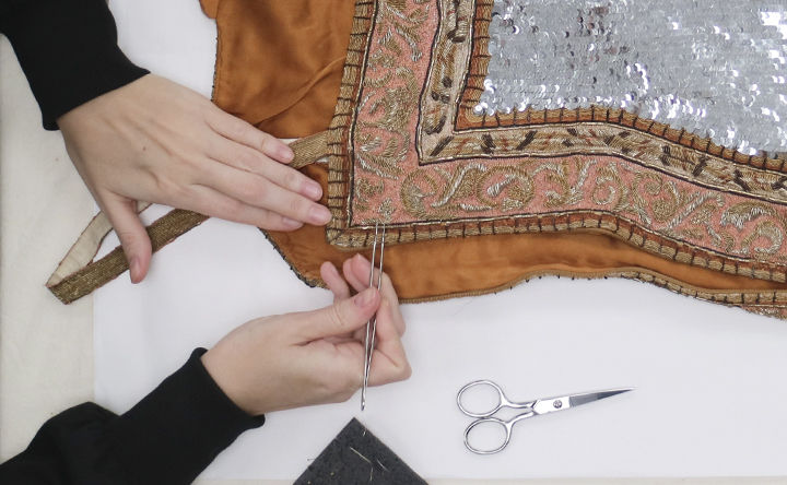 Overhead view of a pair of hands working on a piece of beaded textile; one hand holds the fabric steady while the other holds a long pair of tweezers over a beaded section