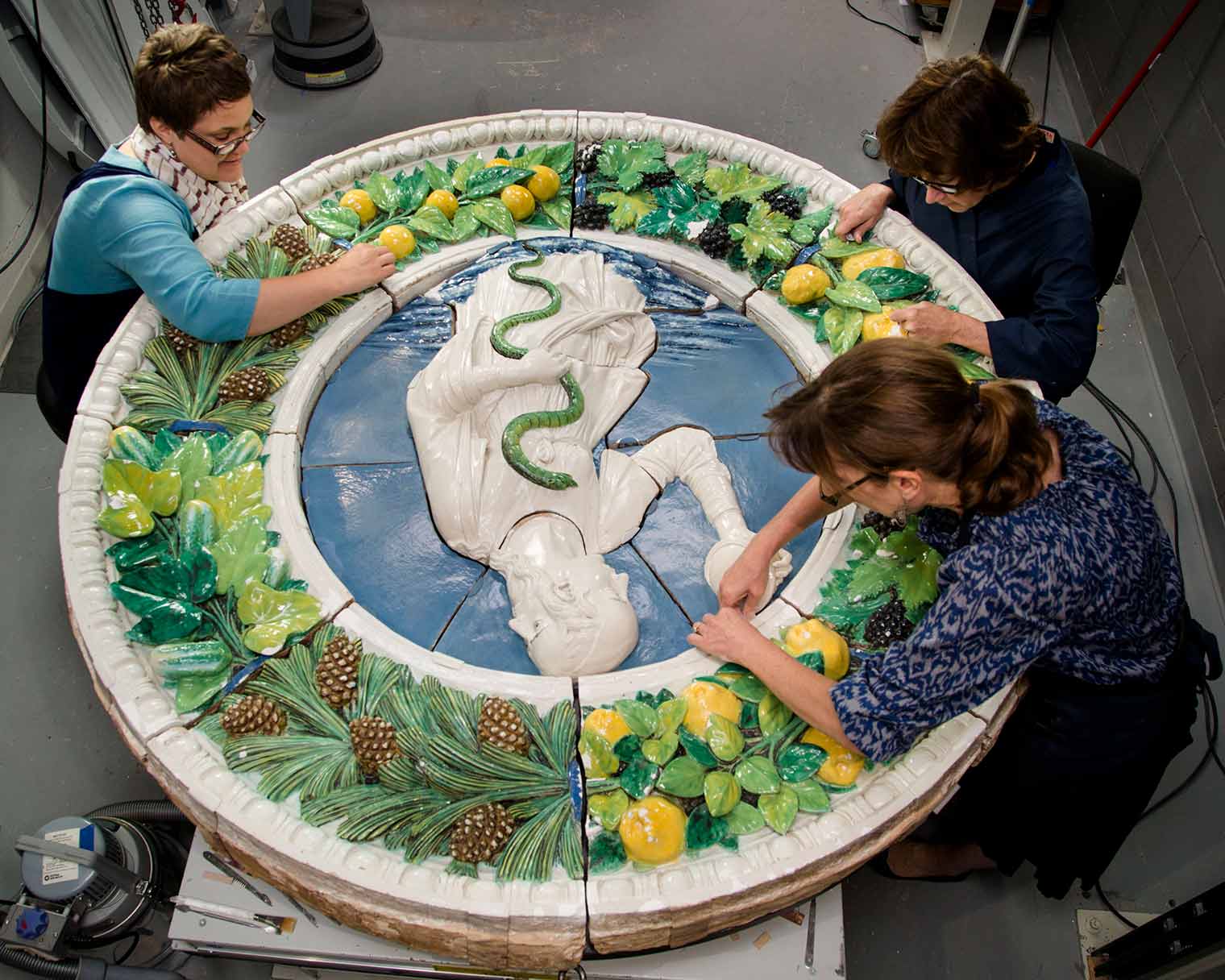 Three conservators working on a large, glazed, ceramic tondo with a human figure with a snake at center
