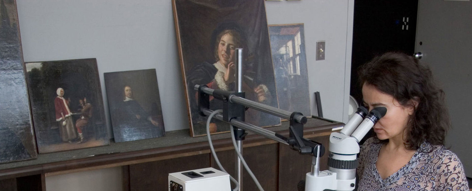 Conservator Isabelle Duvernois examining a Monet landscape through a microscope in the Paintings Conservation studio. Unframed paintings are leaning against a wall on a ledge in the background.
