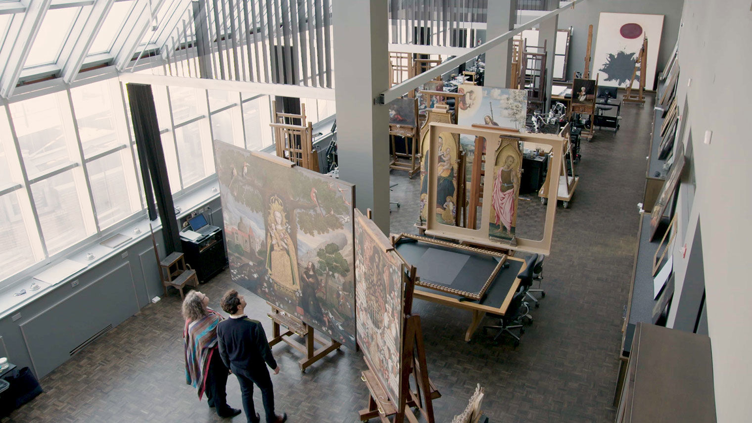 An overhead view of a tall, pitched ceiling room illuminated by a floor-to-ceiling wall of windows and skylights. People work on paintings at several easels placed perpendicularly to the wall of windows.