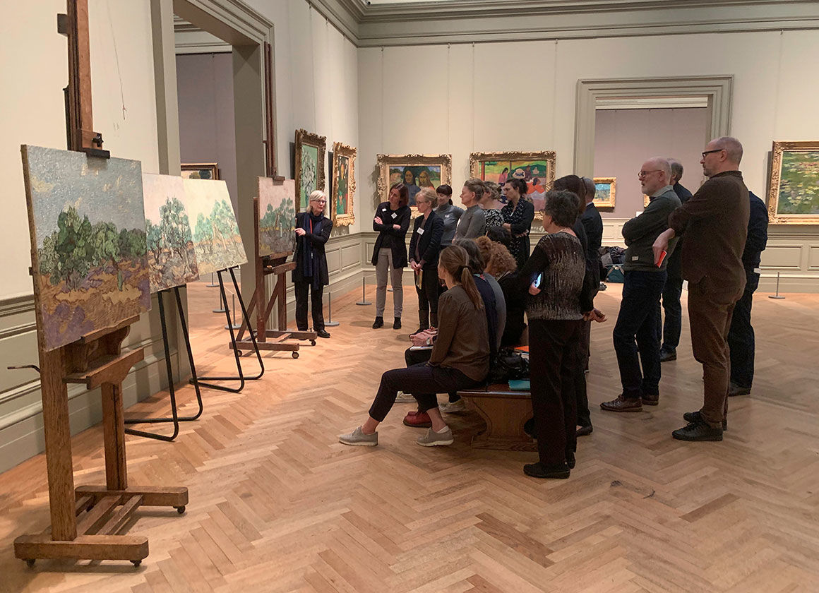 A large group of men and women looking at four paintings on easels stationed in a Museum gallery. A conservator stands at the left of one of the paintings making a presentation to the crowd. The oak floor is set in a herringbone pattern and the walls are white with gray trim.