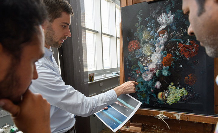 A person holds a print-out and points to a detail of a floral still life mounted on an easel while two other people look on intently. 