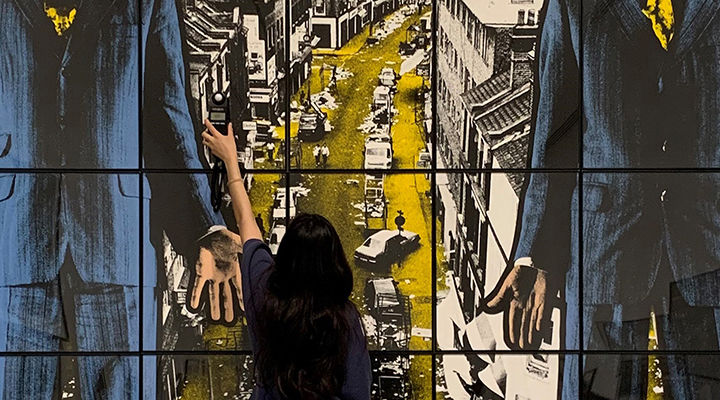 Woman facing large grid of color photographs, reaching up with small light measuring tool.