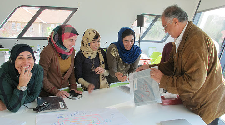 An individual appears to set down and distribute paper resources in plastic sleeves to a group of participants gathered round a white table. The table includes a large marked up sheet of white paper from a brainstorm or teaching session.