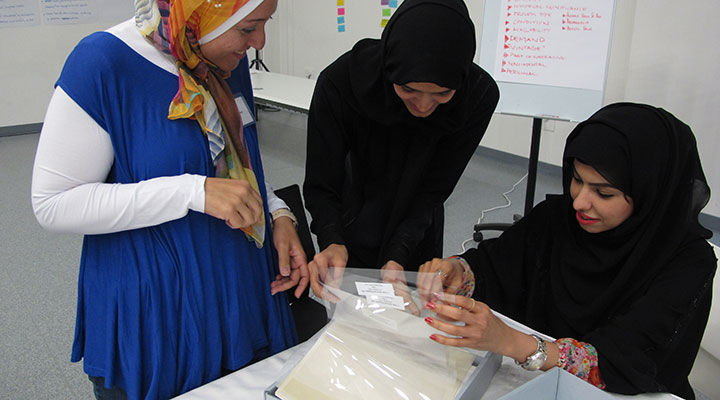 A group of women gather round a table to leaf through and consult papers protected in plastic sleeves stored in archival boxes