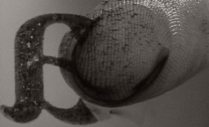 A close-up black-and-white image of a finger smudging an inky lower-case letter "a" off of a transparent surface. The finger tip is so close we see the ridges and lines of the fingerprint.