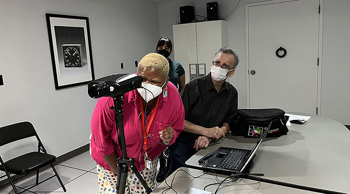 In a bright white room, a young woman, dressed in bright pink, with two colleagues nearby, is leaning forward and looking through a piece of photography equipment called spectroradiometer.