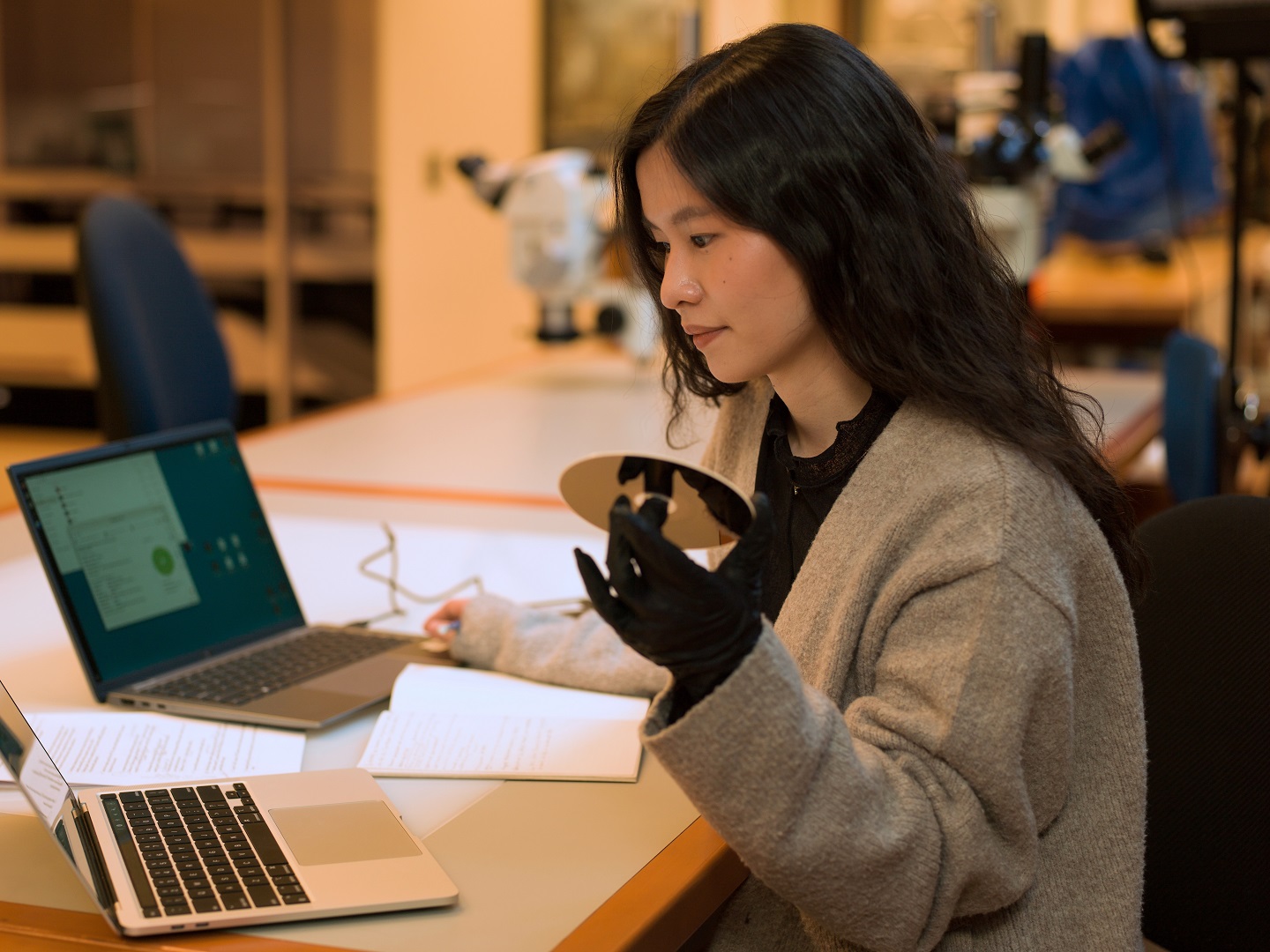A young woman sitting at a work table, wearing nitrile gloves, holding a CD, and working on a laptop.