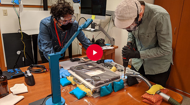 Two conservators wearing clear goggles and masks stand over a large piece of electronic equipment laid out on a large table. One of the conservators holds down the object while the other uses a hand drill to drill or screw into the object. A white triangular play-button set with a red circle appears atop the center of the full image.