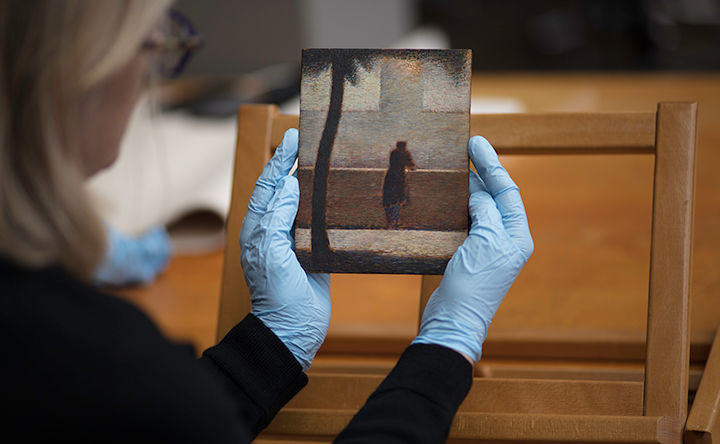 View from over the shoulder of a conservator holding a small rectangular painting in blue rubber gloves; the painting is rendered in short brush strokes that form a kenetic image of a dark, silhouetted tree trunk in the foreground and lone silhouetted figure standing and leaning against a solid, waist-high wall overlooking a faint building in the distance.