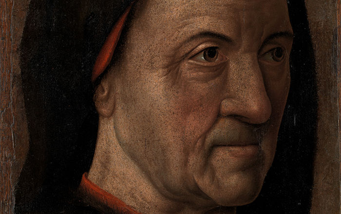 A detail of a portrait by Hugo van der Goes depicting a wrinkled old man wearing a black cloak with a red garment underneath in front of a uniform brown background.