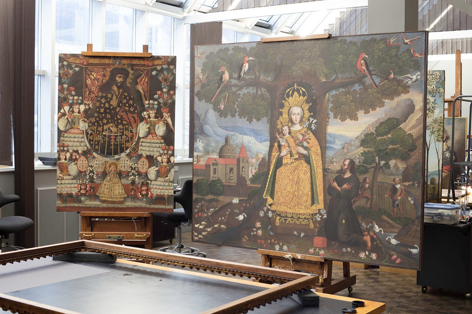 A photograph of two paintings perched on easels within a conservation studio with large windows in the background; the first smaller, portrait-oriented painting on the right depicts a large male figure holding a cross and encircled by a group of five ornately dressed smaller figures; the landscape view painting on the right features a female figure holding white lilies and a baby; both the female figure and child wear ornate crowns and robes; they are set agains a symmetrical background of a tree with tropical-like red birds and a mountaineous landscape; there is a male figure kneeling at the female figure's feet on her right-hand side.