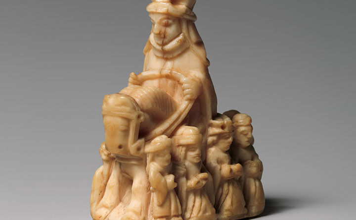 Detail of a small sculptural chess piece made of ivory depicting a large figure riding a horse and surrounded by a group of smaller standing figures.