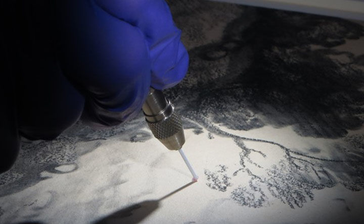 A rubber gloved hand holds and touches a stylus-looking instrument with a long precise tip to a black and white print or drawing of a landscape