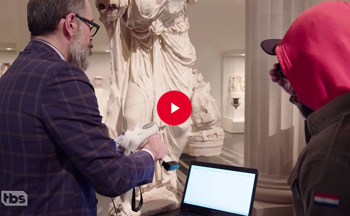 Two individuals study a laptop screen placed in front of a sculptural artwork in the museum galleries