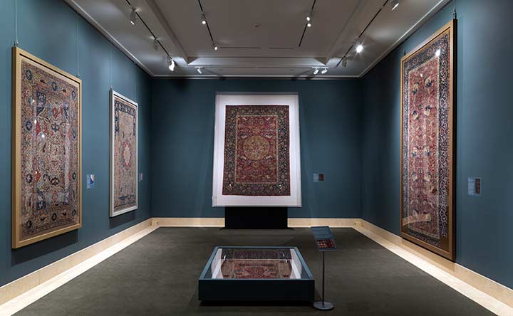 Installation view of "Carpets for Kings: Six Masterpieces of Iranian Weaving" with four large-scale carpets hung on dark teal walls in a small room.
