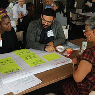 Several seated clusters of people discuss together at tables covered with large note paper and post-its as well as refreshments.