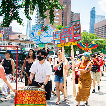 CPP Artist in Residence Mei Lum marches through the streets of Chinatown with event participants holding up large signs of colorfully painted cut-out illustrations of birds and building facades.