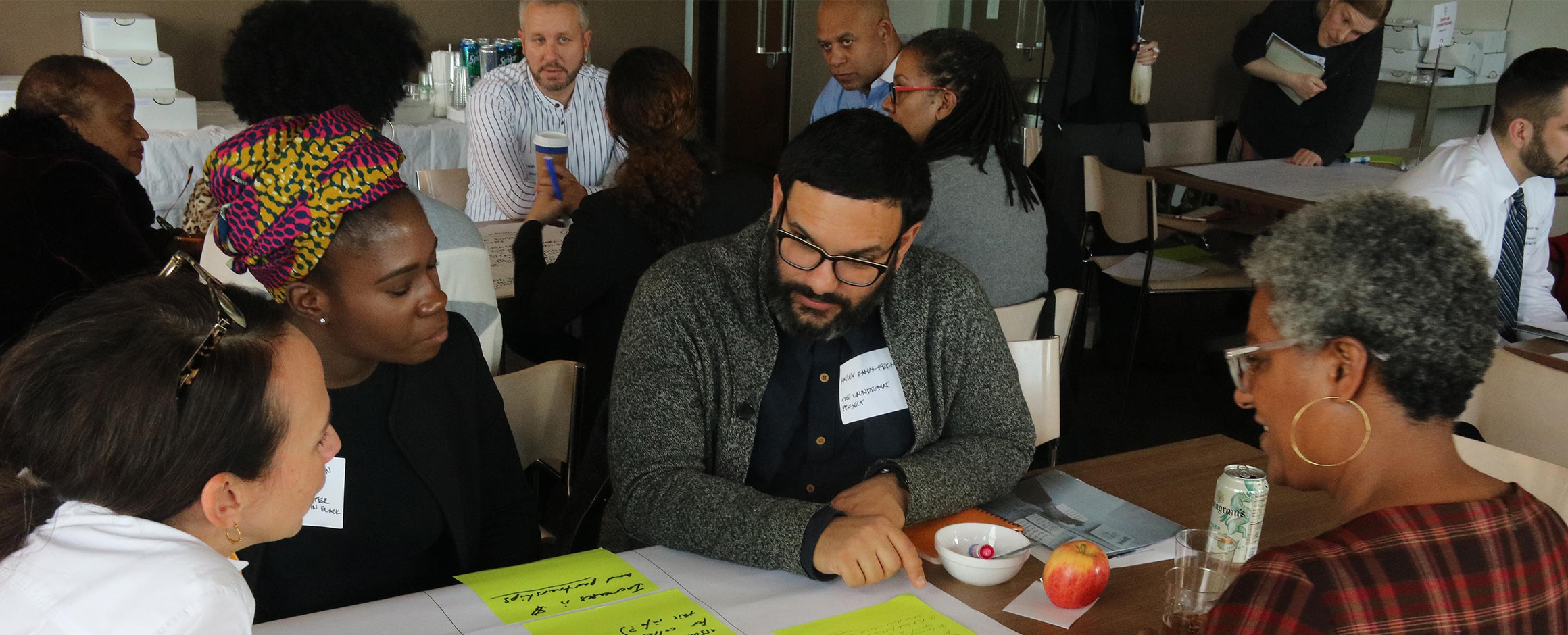 Several seated clusters of people discuss together at tables covered with large note paper and post-its as well as refreshments.