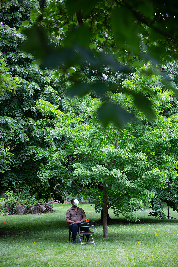 In a grassy area surrounded by trees, a performer sits with their face covered by a silver mask. Another chair faces them.