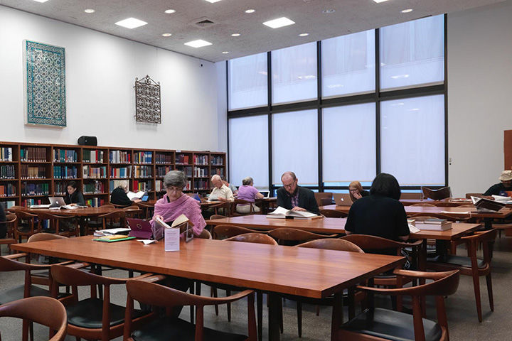 A large, brightly lit room in a library lined with shelves of books; the center has long rectangular wooden tables and chairs where researchers work.