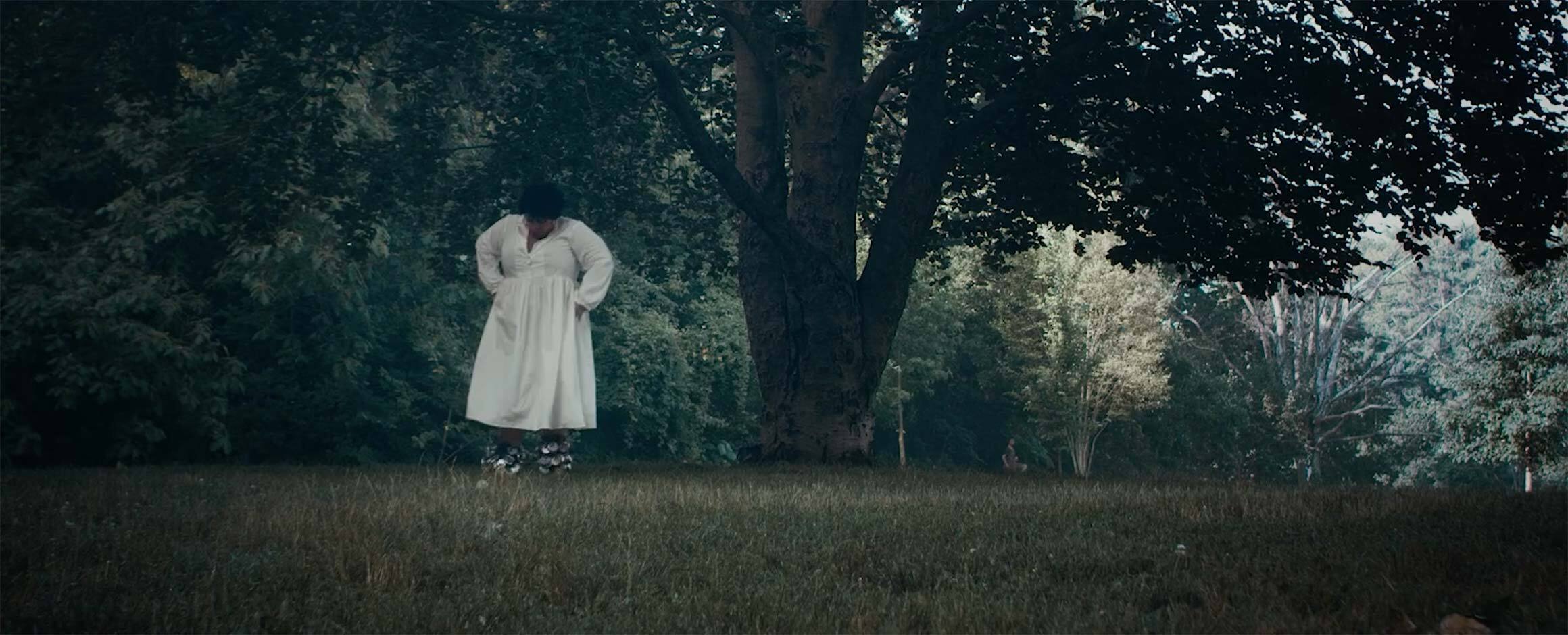 Film still from Rashida Bumbray’s film titled “Braiding and Singing (a point)”; in the film still, Rashida Bumbray wears a white dress, standing in the grass under a large tree in Prospect Park.