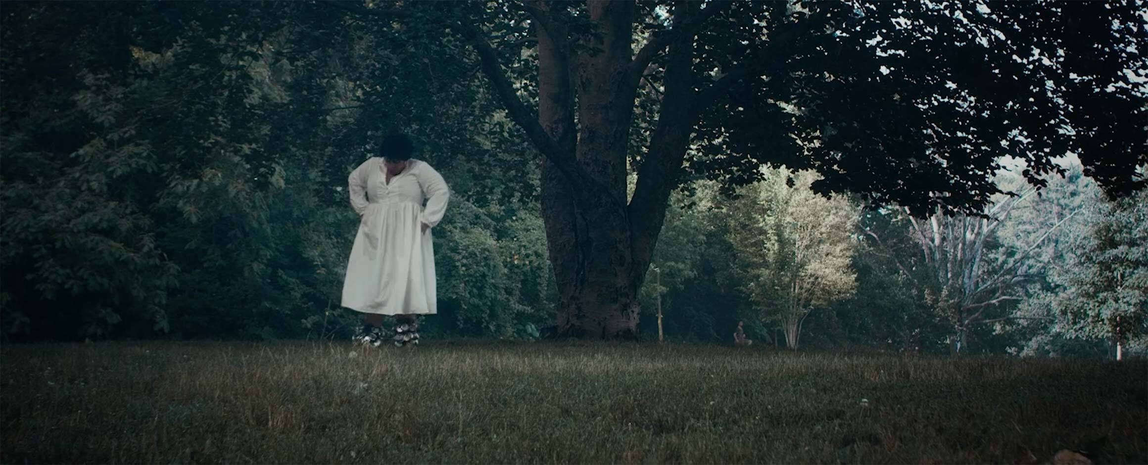 Film still from Rashida Bumbray’s film titled “Braiding and Singing (a point)”; in the film still, Rashida Bumbray wears a white dress, standing in the grass under a large tree in Prospect Park.
