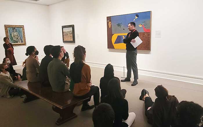 A fellow standing before a modern artwork which is hanging behind him, gives an educational talk in a modern art gallery