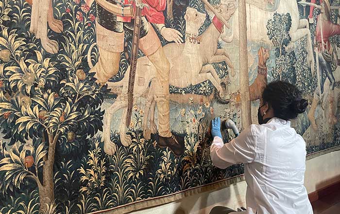 A conservator wearing a white lab coat, and blue protective gloves is mending a delicate and antique textile hanging on a wall.