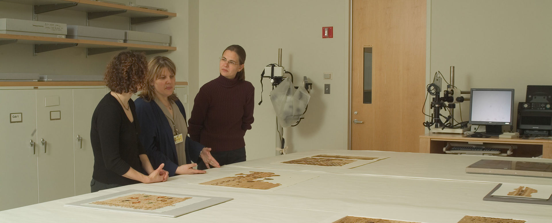 Three woman stand side by side in front of a large white table splayed out with textiles