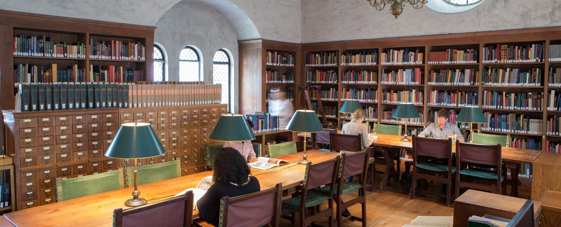 A small library lined with dark wood book shelves and tables with lamps and green upholstered chairs, arched windows, and a stained glass rosette set high in the far wall