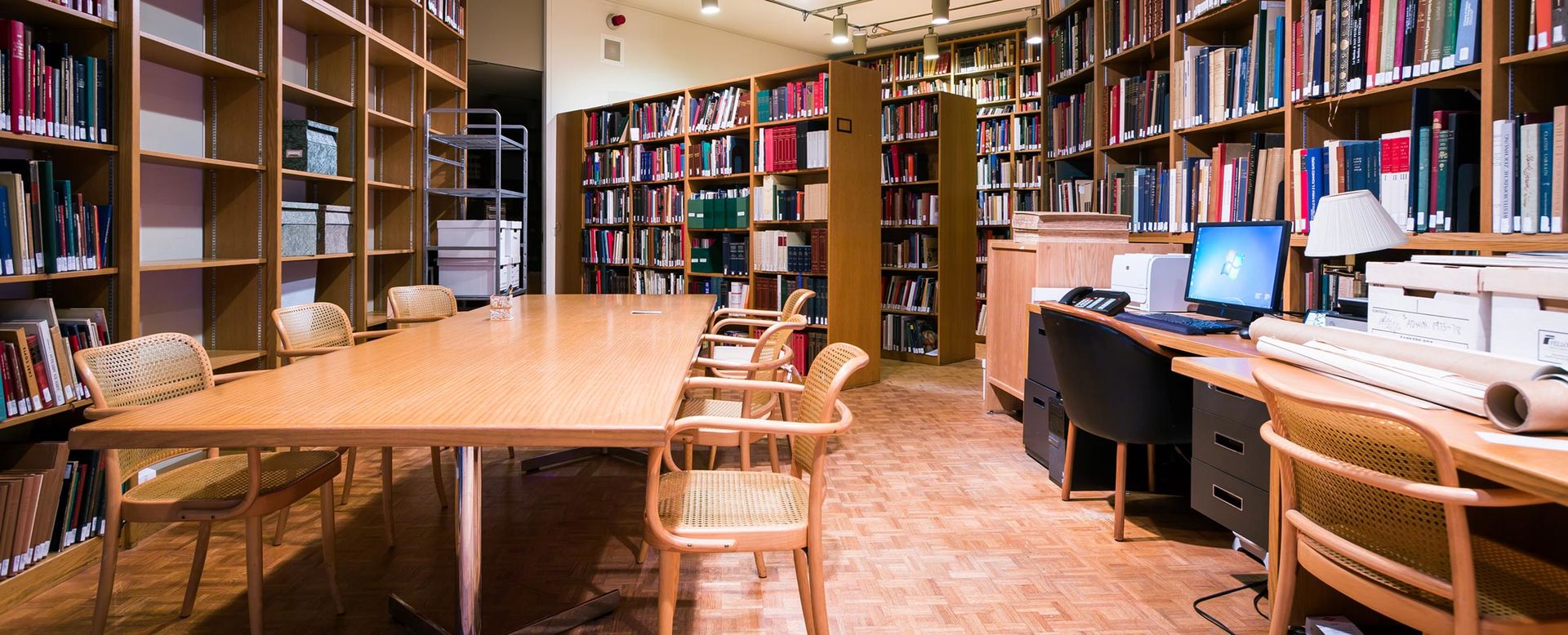 A narrow modern library with blonde wood furniture and lined with shelves of books; through an archway, another room lined with books and bookshelves can be seen