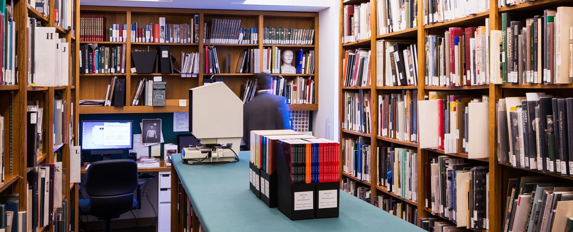 A small, brightly lit library filled with shelves of books with a man looking at the back shelves