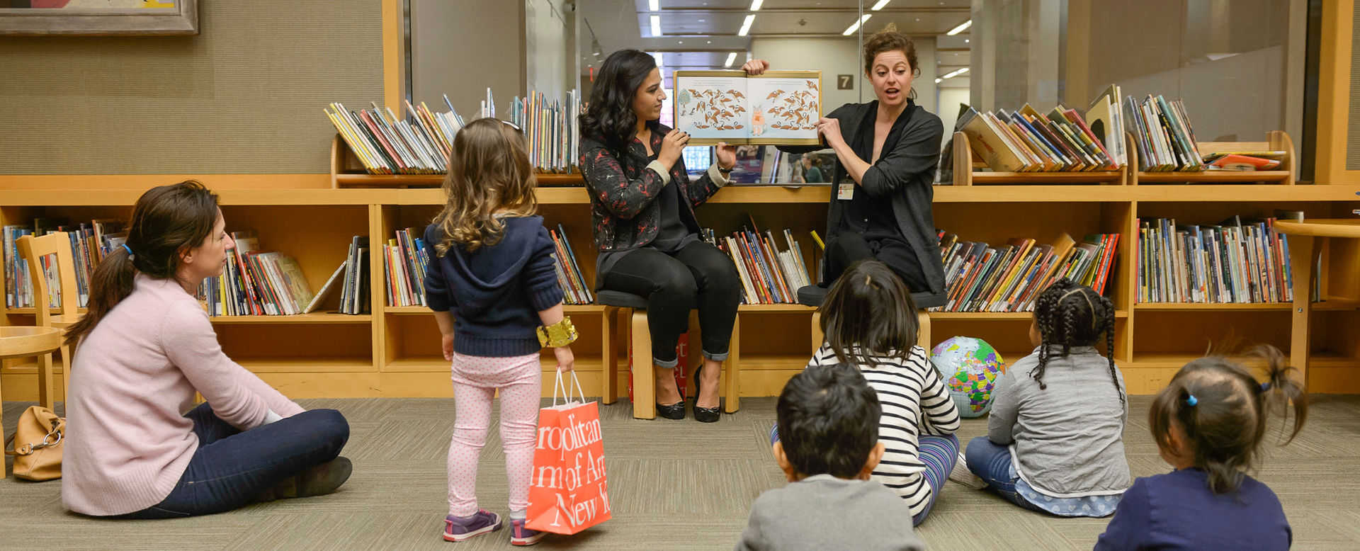 Two women display a picture book as they read to five children in a modern, well-lit, spacious, and open library with low bookshelves filled with books