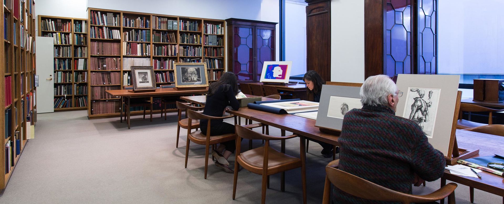 A narrow library with a long dark wood table and glass fronted cabinets with a dark-haired woman and elderly man looking at art, and in the background shelves of books