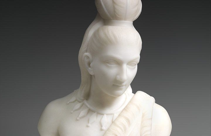 alt text="A white marble bust of a Native American male with feather headdress and necklace"