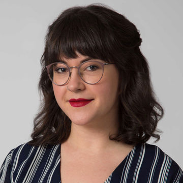 A smiling woman with long dark hair and glasses, wearing a black top with thin white vertical stripes