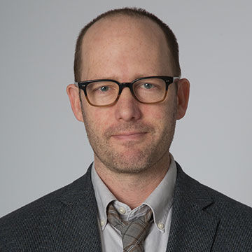 A man with close cropped dark hair and glasses, wearing a dark gray jacket over an open collared gray shirt and tie