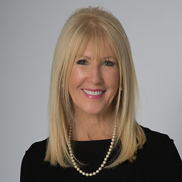 A woman with long blonde hair, wearing a black blouse with pearls